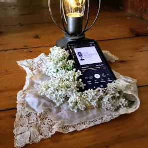 A smartphone rests on a wooden surface and displays a podcast episode, surrounded by a lace cloth and white lilac flowers. A decorative lantern with a glowing light bulb stands nearby.
