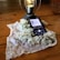 A smartphone rests on a wooden surface and displays a podcast episode, surrounded by a lace cloth and white lilac flowers. A decorative lantern with a glowing light bulb stands nearby.