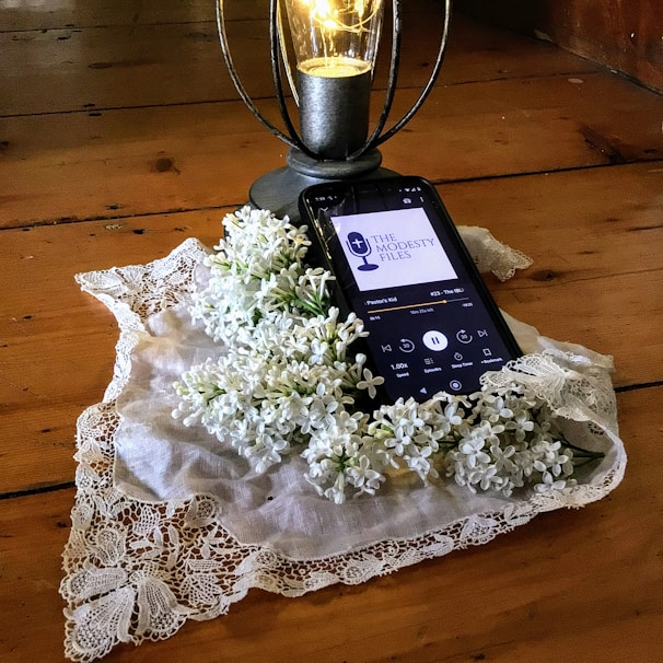 A smartphone rests on a wooden surface and displays a podcast episode, surrounded by a lace cloth and white lilac flowers. A decorative lantern with a glowing light bulb stands nearby.
