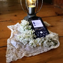 A smartphone rests on a wooden surface and displays a podcast episode, surrounded by a lace cloth and white lilac flowers. A decorative lantern with a glowing light bulb stands nearby.