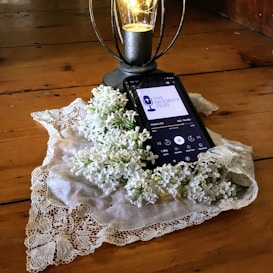 A smartphone rests on a wooden surface and displays a podcast episode, surrounded by a lace cloth and white lilac flowers. A decorative lantern with a glowing light bulb stands nearby.