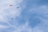Three parachutists are descending against a bright blue sky scattered with wispy clouds. The parachutes are vibrant in color, with two being red and one being a combination of green and blue. The scene conveys a sense of freedom and adventure.