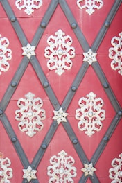 Intricate metallic decorations with symmetrical, floral patterns are arranged in a diamond lattice design on a red background. Dark metal strips form the diamond shapes, intersected by lighter ornamental pieces in a symmetrical pattern.