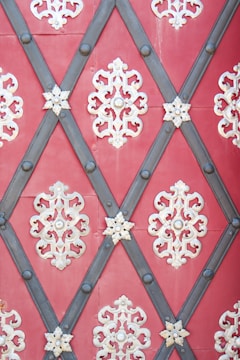 Intricate metallic decorations with symmetrical, floral patterns are arranged in a diamond lattice design on a red background. Dark metal strips form the diamond shapes, intersected by lighter ornamental pieces in a symmetrical pattern.