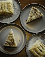 Four dessert plates with pastries, including triangular slices with sprinkle toppings and rectangular pastries dusted with sugar, arranged on a dark wooden table.