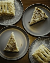Four dessert plates with pastries, including triangular slices with sprinkle toppings and rectangular pastries dusted with sugar, arranged on a dark wooden table.