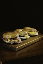 Three bagels are placed on a wooden board, filled with bacon and cheese, set against a dark background.