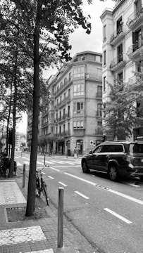 A grayscale image of a city street featuring several multi-story buildings with balconies and ornate architecture. The street is lined with trees and has a bike leaning against a pole. A pedestrian is crossing at a crosswalk, and a large SUV is stopped at a traffic light. The sidewalks have distinct paving patterns and there is minimal pedestrian activity.