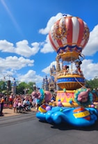 A festive parade float shaped like a hot air balloon is moving through a theme park. The float is vibrantly decorated with red, white, and blue stripes, adorned with colorful patterns and balloons. It features popular cartoon characters standing on the basket of the hot air balloon. In the background, there is a large castle with blue rooftops, surrounded by lush greenery and a clear blue sky with fluffy clouds. A large crowd of spectators lines the street, capturing photos and enjoying the parade.