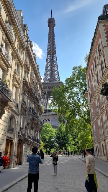 A street view leading to the Eiffel Tower, framed by elegant Parisian buildings with ornate balconies on both sides. The area is lined with lush green trees under a clear blue sky. People are casually walking and taking photos along the cobblestone street.