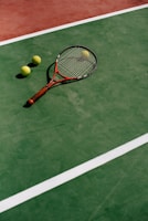 A tennis racket with a brown handle and three yellow tennis balls are positioned on a green tennis court, marked with white lines. The surface of the court has a slightly worn texture, indicating usage.