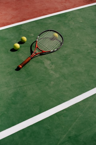 A tennis racket with a brown handle and three yellow tennis balls are positioned on a green tennis court, marked with white lines. The surface of the court has a slightly worn texture, indicating usage.
