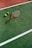 A tennis racket with a brown handle and three yellow tennis balls are positioned on a green tennis court, marked with white lines. The surface of the court has a slightly worn texture, indicating usage.