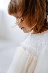A young child with light brown hair is captured in profile. The child is wearing a white dress with floral embroidery, appearing soft and delicate. Light softly illuminates the child's face, creating a serene and peaceful atmosphere.