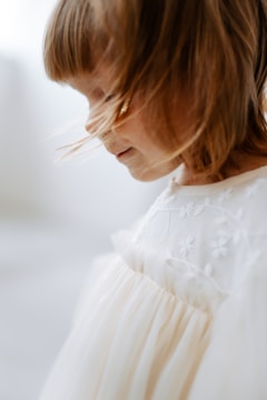A young child with light brown hair is captured in profile. The child is wearing a white dress with floral embroidery, appearing soft and delicate. Light softly illuminates the child's face, creating a serene and peaceful atmosphere.