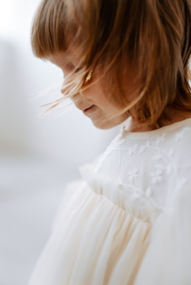 A young child with light brown hair is captured in profile. The child is wearing a white dress with floral embroidery, appearing soft and delicate. Light softly illuminates the child's face, creating a serene and peaceful atmosphere.