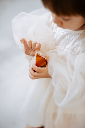 A young child wearing an ornate white dress with floral embroidery is delicately holding a partially unwrapped muffin. The soft fabric of the dress contrasts with the rich orange color of the muffin.