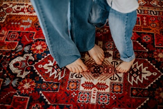 Two pairs of bare feet are visible on a vibrant, intricately patterned Persian-style rug. The individuals are wearing blue jeans, and their feet are placed closely together, suggesting a sense of intimacy or connection. The rug features a rich design with various geometric and floral patterns.