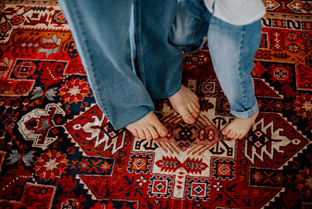 Two pairs of bare feet are visible on a vibrant, intricately patterned Persian-style rug. The individuals are wearing blue jeans, and their feet are placed closely together, suggesting a sense of intimacy or connection. The rug features a rich design with various geometric and floral patterns.