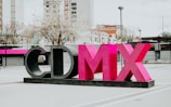A large sculptural installation featuring the letters 'CDMX' in bold, vibrant pink and black colors stands prominently in an urban outdoor setting. Surrounding the structure are modern buildings, leafless trees, and a lamppost, contributing to a cityscape environment.