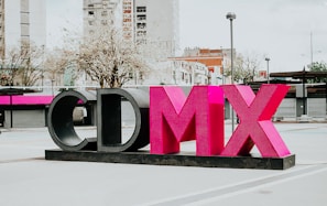 A large sculptural installation featuring the letters 'CDMX' in bold, vibrant pink and black colors stands prominently in an urban outdoor setting. Surrounding the structure are modern buildings, leafless trees, and a lamppost, contributing to a cityscape environment.
