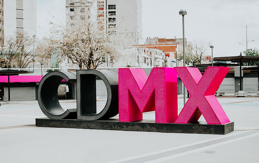 A large sculptural installation featuring the letters 'CDMX' in bold, vibrant pink and black colors stands prominently in an urban outdoor setting. Surrounding the structure are modern buildings, leafless trees, and a lamppost, contributing to a cityscape environment.