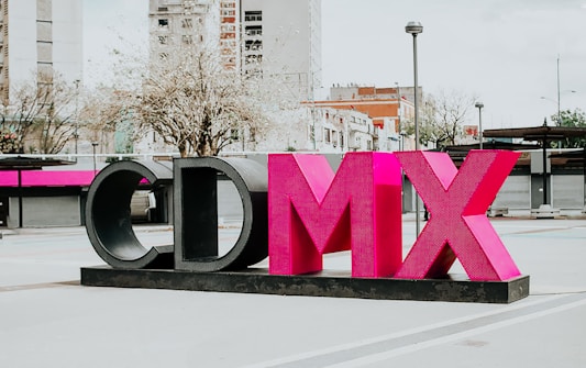 A large sculptural installation featuring the letters 'CDMX' in bold, vibrant pink and black colors stands prominently in an urban outdoor setting. Surrounding the structure are modern buildings, leafless trees, and a lamppost, contributing to a cityscape environment.