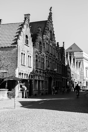 A black-and-white photograph of a historic European street with ornate, gabled buildings made of brick. The cobblestone street is occupied by pedestrians and a cyclist. Shadows are cast across the pavement, conveying a sense of late afternoon or early morning light.