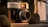 A vintage camera with the brand name 'Voigtlander' is prominently displayed in a close-up shot. The camera's intricate details and metal components are highlighted by warm, golden lighting. Surrounding the camera are some green leaves, which add a natural element to the composition.