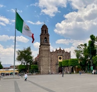 A tall, historic stone building with a bell tower stands prominently. The large Mexican flag flutters in the wind on a flagpole. The setting is a spacious plaza with a few people walking around and trees lining the area. The sky is partly cloudy, casting a bright and lively atmosphere.