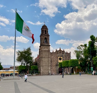 A tall, historic stone building with a bell tower stands prominently. The large Mexican flag flutters in the wind on a flagpole. The setting is a spacious plaza with a few people walking around and trees lining the area. The sky is partly cloudy, casting a bright and lively atmosphere.