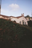 A charming white house with a red-tiled roof sits partially obscured by lush greenery. The architecture features arched windows and a chimney, giving a classic Mediterranean vibe. In the background, there is a clear blue sky with a few clouds.