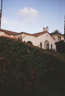 A charming white house with a red-tiled roof sits partially obscured by lush greenery. The architecture features arched windows and a chimney, giving a classic Mediterranean vibe. In the background, there is a clear blue sky with a few clouds.
