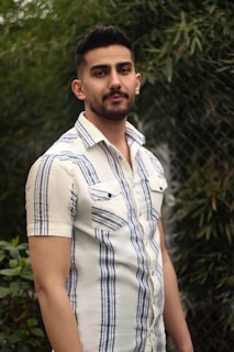 A man with short, dark hair and a trimmed beard is wearing a short-sleeved, button-up shirt with blue and white vertical stripes. He is standing outdoors in front of a green backdrop with plants and a chain-link fence, giving a relaxed and natural setting.