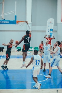 A basketball game in progress with players actively engaging on the court. One player in a black and red jersey is performing a slam dunk while others in blue and white jerseys observe or attempt to block. The court is well-lit, and there are banners and seating visible in the background.