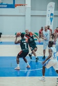 A basketball game is in progress with players in action near the hoop on a court. One player in a dark uniform is attempting a pass or a shot, while a player in a white uniform is closely guarding him. Other players in both dark and white uniforms can be seen in the background observing or preparing to move.