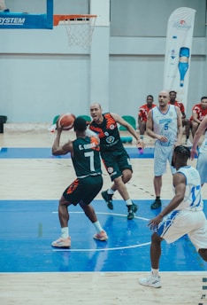 A basketball game is in progress with players in action near the hoop on a court. One player in a dark uniform is attempting a pass or a shot, while a player in a white uniform is closely guarding him. Other players in both dark and white uniforms can be seen in the background observing or preparing to move.
