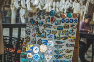 A display board is filled with a variety of colorful souvenir magnets and trinkets. There are numerous designs, including evil eye symbols, maps, and animal shapes, all with a theme related to Cyprus. In the background, a person is sitting at a table, slightly out of focus, and there are soft white feathery decorations hanging from above.