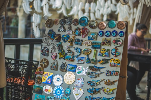A display board is filled with a variety of colorful souvenir magnets and trinkets. There are numerous designs, including evil eye symbols, maps, and animal shapes, all with a theme related to Cyprus. In the background, a person is sitting at a table, slightly out of focus, and there are soft white feathery decorations hanging from above.