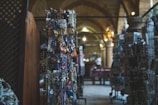 A dimly lit interior of a market or bazaar with displays of various colorful trinkets and souvenirs hanging from racks. The background features stone arches and soft lighting, adding a rustic ambiance.
