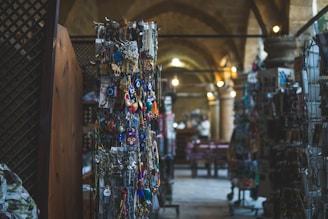 A dimly lit interior of a market or bazaar with displays of various colorful trinkets and souvenirs hanging from racks. The background features stone arches and soft lighting, adding a rustic ambiance.
