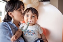 A woman is gently kissing a baby's cheek, who is wearing a pacifier with a green clip attached to their clothing. The baby is dressed in a t-shirt with a floral pattern and the word 'flower' printed on it. The background contains softly blurred, neutral colors, creating a warm and intimate atmosphere.