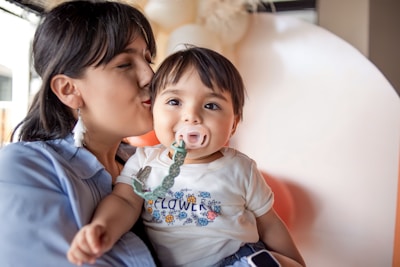 A woman is gently kissing a baby's cheek, who is wearing a pacifier with a green clip attached to their clothing. The baby is dressed in a t-shirt with a floral pattern and the word 'flower' printed on it. The background contains softly blurred, neutral colors, creating a warm and intimate atmosphere.