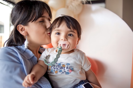 A woman is gently kissing a baby's cheek, who is wearing a pacifier with a green clip attached to their clothing. The baby is dressed in a t-shirt with a floral pattern and the word 'flower' printed on it. The background contains softly blurred, neutral colors, creating a warm and intimate atmosphere.