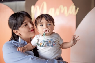 A woman is lovingly holding a baby who has a pacifier in their mouth. The baby is wearing a white shirt with colorful floral designs and the word 'Flower' printed on it. The background features a soft, peachy tone with the word 'Mamá' visible.
