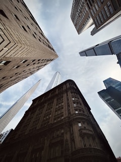 A dramatic upward view of a cluster of towering urban skyscrapers, emphasizing the architectural diversity and grandeur of city buildings. The sky is partially visible through the narrow gaps left by the converging facades above.