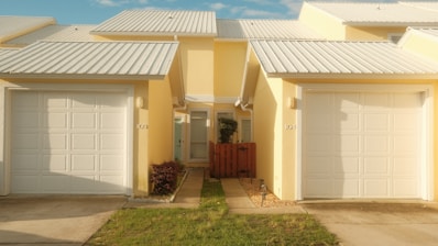 Side-by-side yellow houses with identical architecture feature white garages and metal roofing. A small patch of grass and shrubs line the pathway to the doors, separated by a wooden fence.