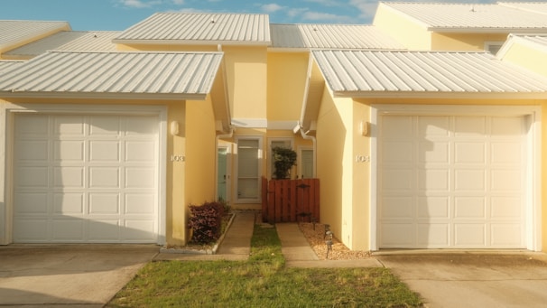 Side-by-side yellow houses with identical architecture feature white garages and metal roofing. A small patch of grass and shrubs line the pathway to the doors, separated by a wooden fence.