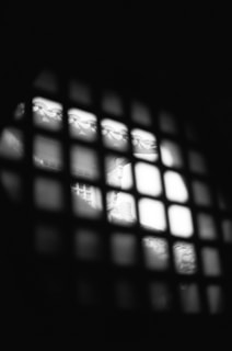 A dimly lit image featuring a grid pattern in the foreground, with bright light illuminating part of a scene behind it. The scene appears blurry but displays elements like intricate patterns and structures.