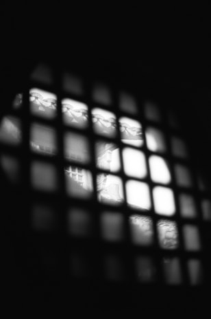 A dimly lit image featuring a grid pattern in the foreground, with bright light illuminating part of a scene behind it. The scene appears blurry but displays elements like intricate patterns and structures.
